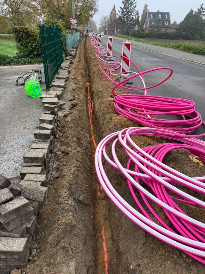 Graben am Straßenrand mit pinken Kabeln und Baufahrzeugen in der Nähe.