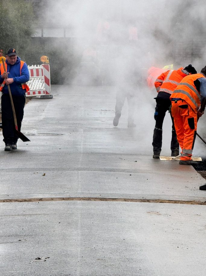 Arbeiter in Warnkleidung reinigen eine nasse Straße, umgeben von Dampf.