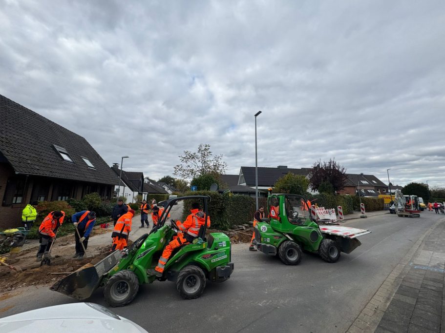 Bauarbeiter mit Maschinen arbeiten an einer Straße in einem Wohngebiet.