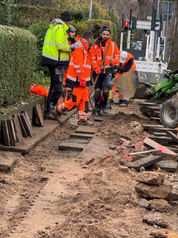 Arbeiter in orangefarbener Kleidung führen Erdarbeiten an einer Straße durch.