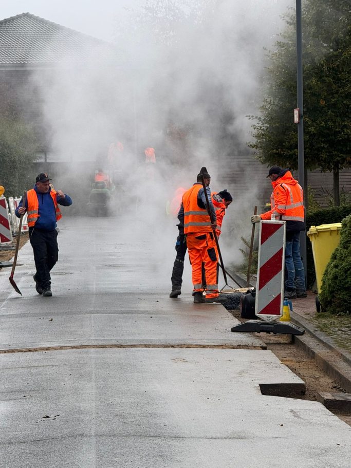 Arbeiter in orangefarbener Kleidung führen Straßenreparaturen durch, umgeben von Dampf.