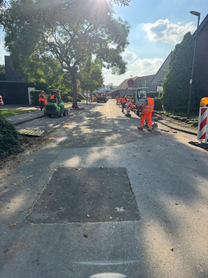 Bauarbeiten auf einer Straße mit Arbeiter in orangefarbener Kleidung und Maschinen.